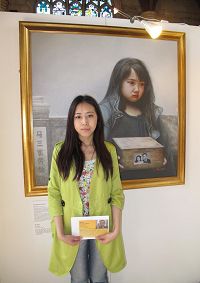 '圖4：於銘慧手裏拿著營救父親的明信片站在「真善忍國際美展」畫作「孤兒淚」前'